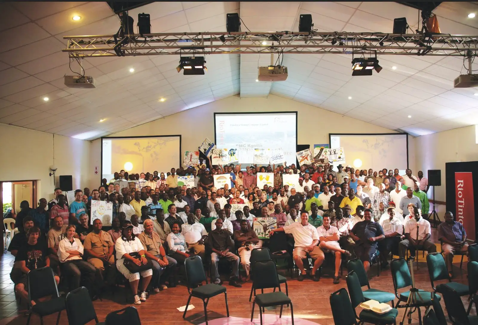 Hundreds of participants seated in a hall facing three screens