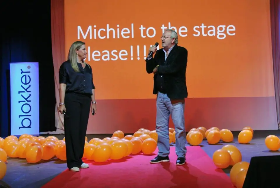 Two presenters on stage under a screen reading “Michiel to the stage please!!!”