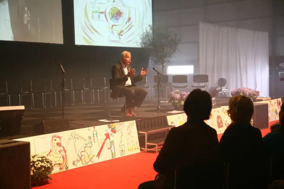 Speaker seated on stage beside a drawing with the Red Cross emblem