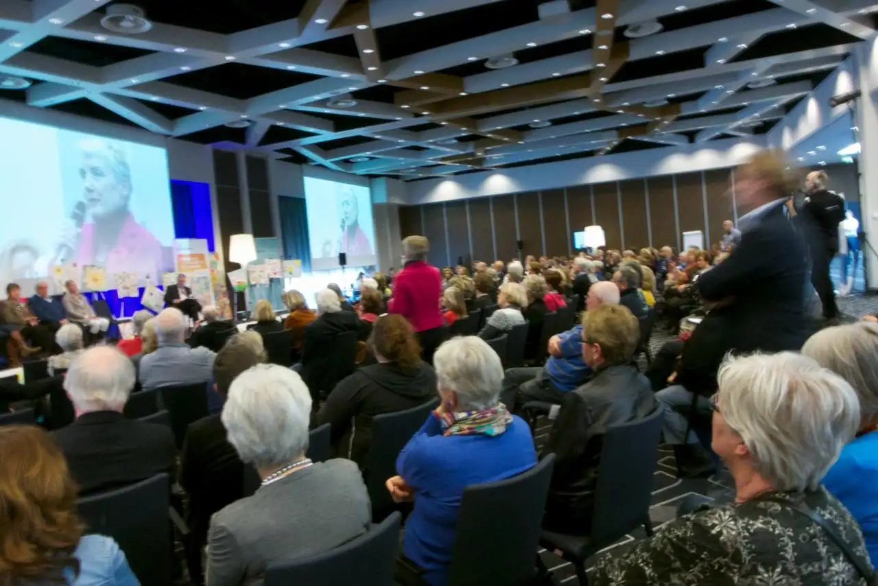 Audience seated in a conference hall facing a speaker on a large screen